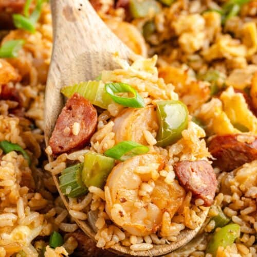 A wooden spoon scooping up cajun fried rice with shrimp, sausage, rice, green bell pepper, celery, and chopped green onions.