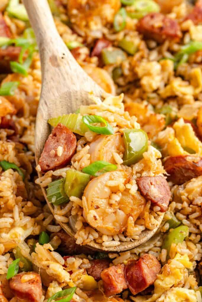 A wooden spoon scooping up cajun fried rice with shrimp, sausage, rice, green bell pepper, celery, and chopped green onions.