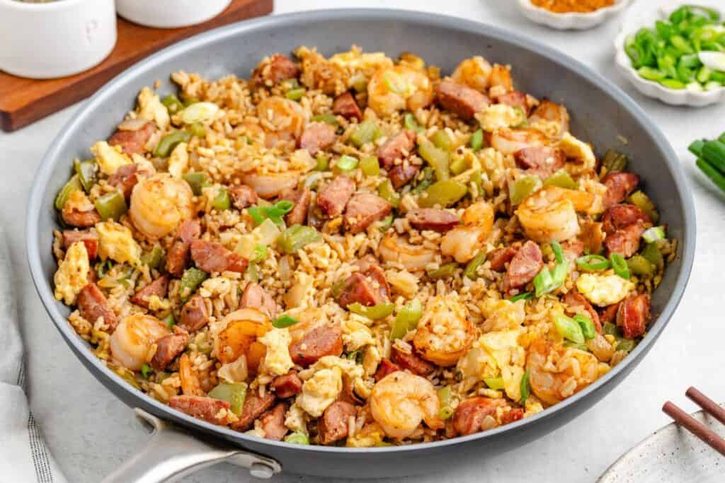 A large skillet filled with cajun fried rice, shrimp, sausage, scrambled eggs, green onions, and chopped green peppers, all mixed together and cooked.