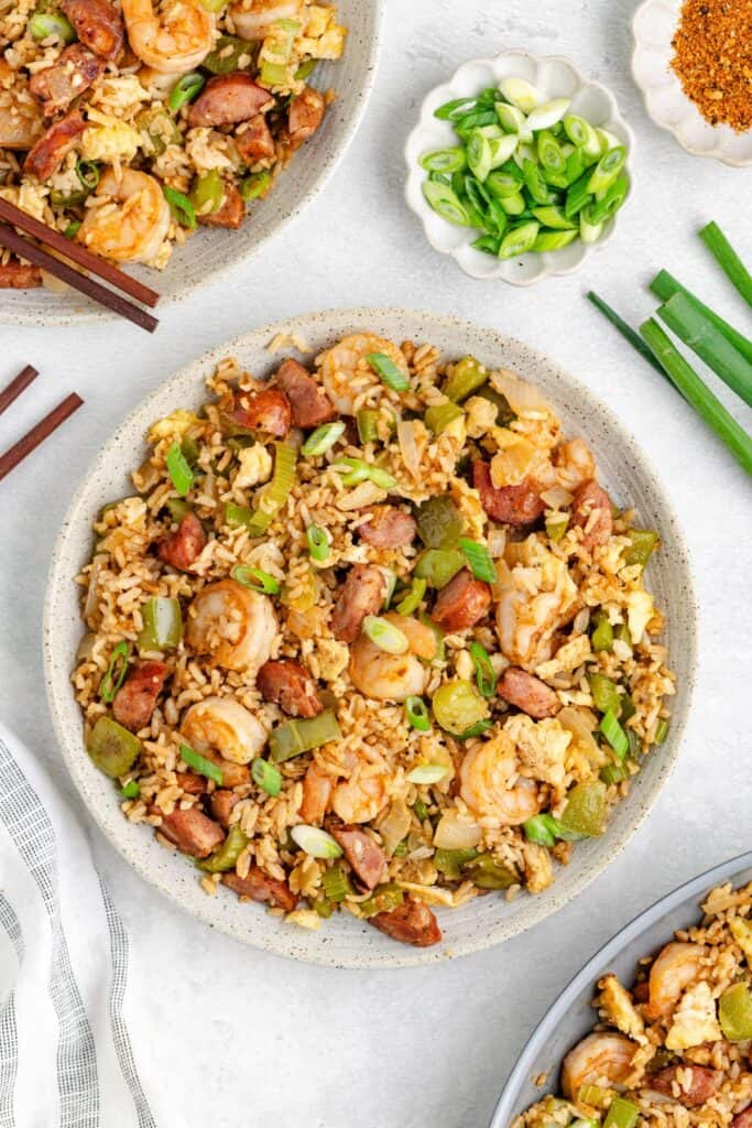 A bowl of cajun fried rice with sausage, scrambled eggs, green onions, and vegetables. Sliced green onions and chopsticks are on the side, with more rice in a partial bowl in the corner.
