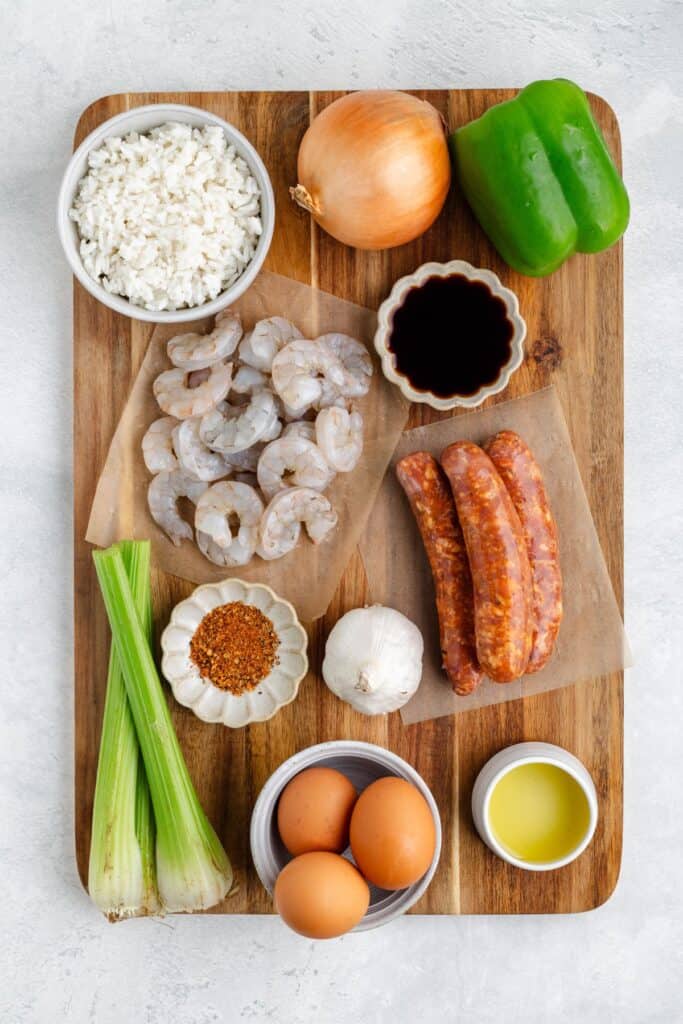 A wooden board displays raw shrimp, sausages, rice, a green bell pepper, an onion, celery stalks, garlic, eggs, soy sauce, oil, and a small dish of red seasoning on a light background.