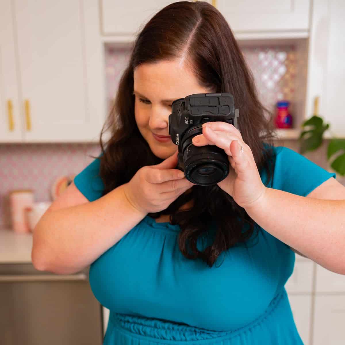 Woman in teal dress photographing with camera in a sunlit kitchen, ready to take a picture.