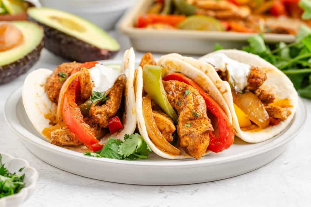 Chicken fajitas in three flour tortillas with sautéed peppers, onions, sour cream, and cilantro on a white plate; avocado halves behind.