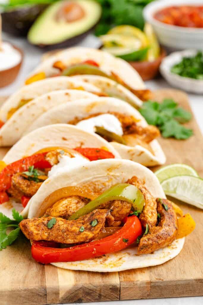 Chicken fajita in tortillas with grilled peppers, onions, cilantro, and lime on a wooden board; toppings behind.