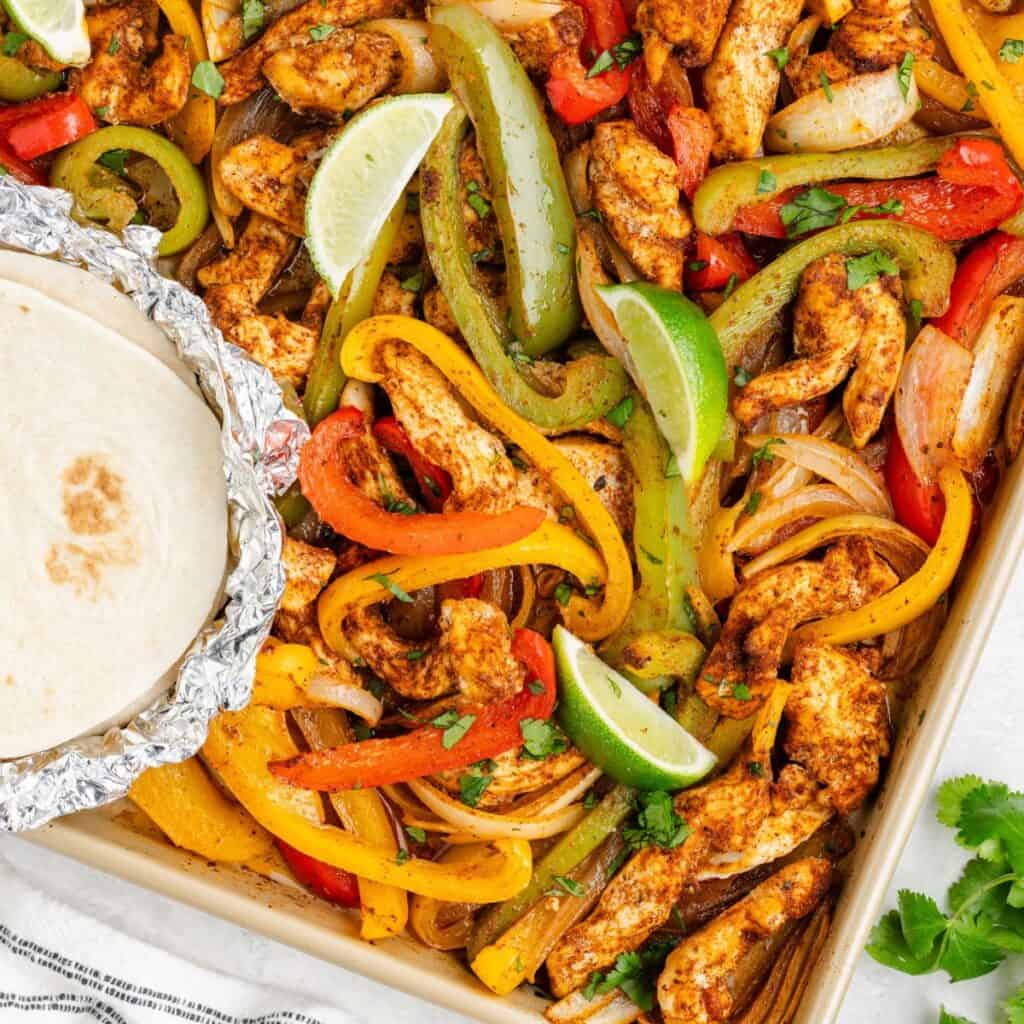 Sheet pan with roasted chicken strips, bell peppers, onions, lime wedges, cilantro, and foil-wrapped tortillas.