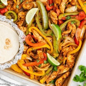Sheet pan with roasted chicken strips, bell peppers, onions, lime wedges, cilantro, and foil-wrapped tortillas.