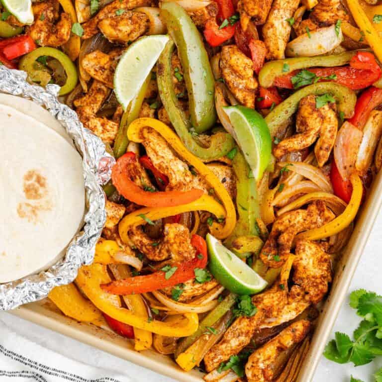 Sheet pan with roasted chicken strips, bell peppers, onions, lime wedges, cilantro, and foil-wrapped tortillas.