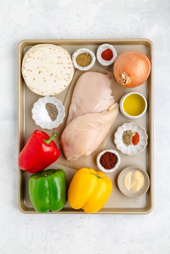 Sheet pan with raw chicken, colorful bell peppers, onion, tortillas, olive oil, and spices for fajitas.