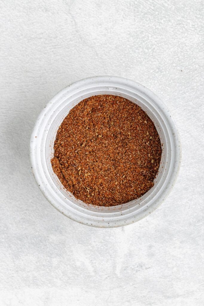 White bowl of reddish-brown spice blend for chicken fajitas on a light gray textured surface.