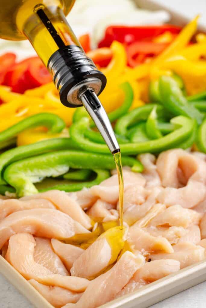 Olive oil poured on raw chicken strips, with sliced red, yellow, and green bell peppers in the background.