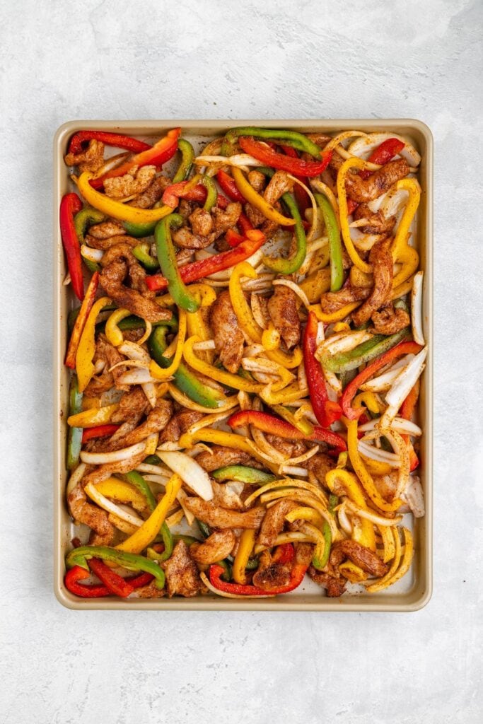 Sheet pan with sliced seasoned chicken, red, yellow, green bell peppers, and onions arranged for baking fajitas.