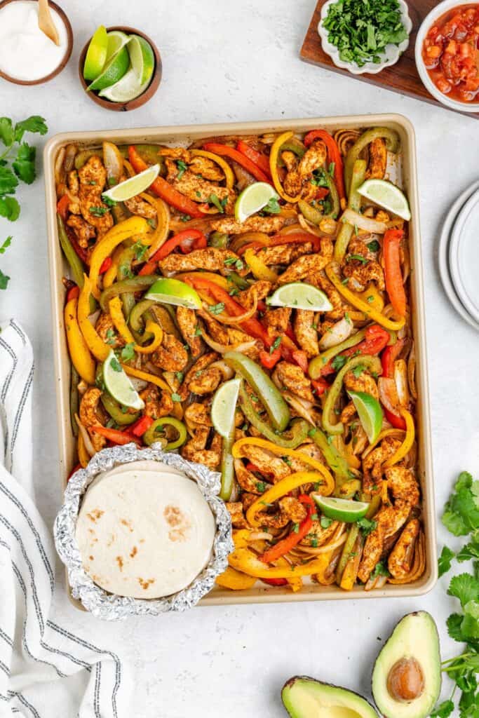 Sheet pan chicken fajitas with peppers and lime, surrounded by tortillas, avocado, cilantro, sour cream, and salsa.