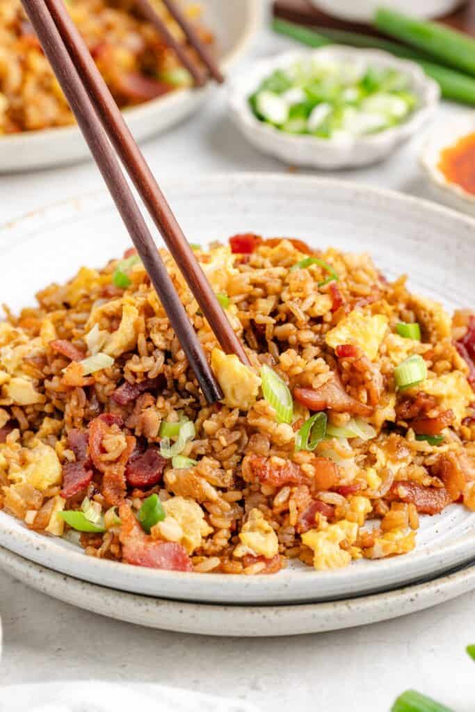 Chopsticks picking up breakfast fried rice with scrambled eggs, green onions, and bacon from a white plate.