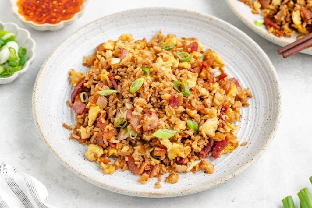 Fried rice for breakfast with scrambled eggs, bacon, and green onions on a white plate; sauce and scallions beside.