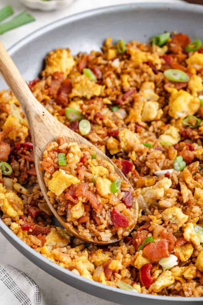 Skillet of breakfast fried rice with eggs, bacon, and green onions; wooden spoon scoops a portion.