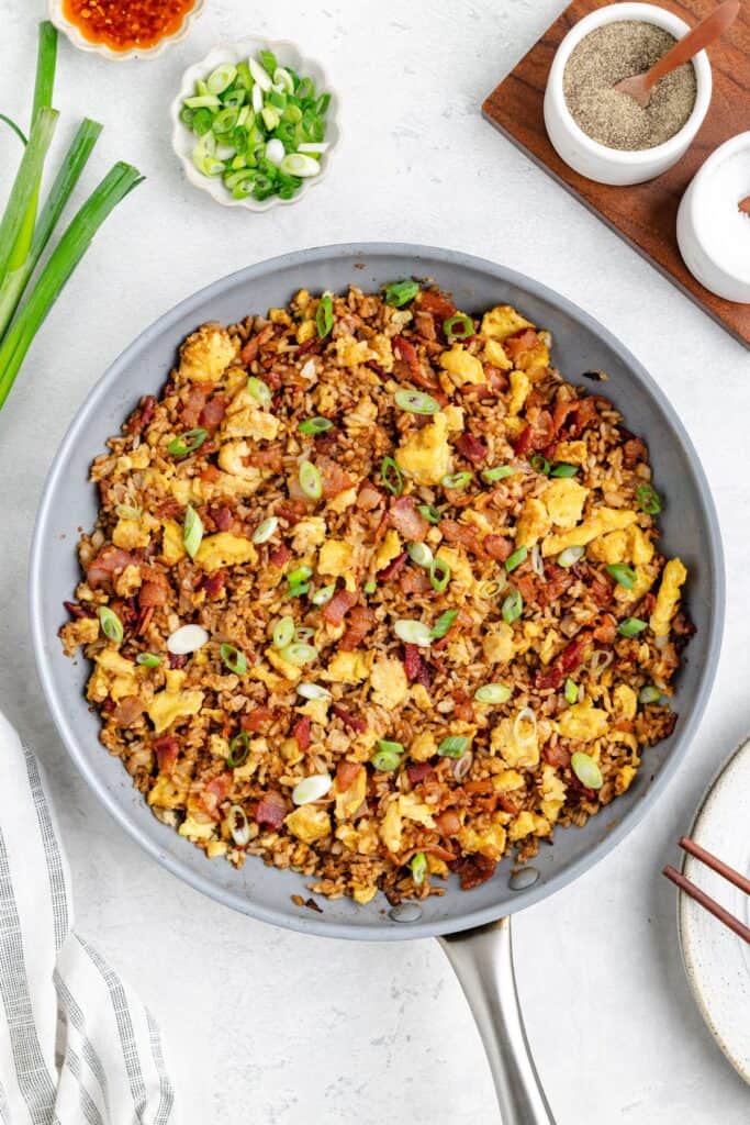 A skillet bacon and egg fried rice with green onions, surrounded by bowls of seasonings.