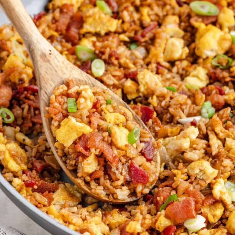 Wooden spoon scooping breakfast fried rice with scrambled eggs, bacon, and green onions from a pan.