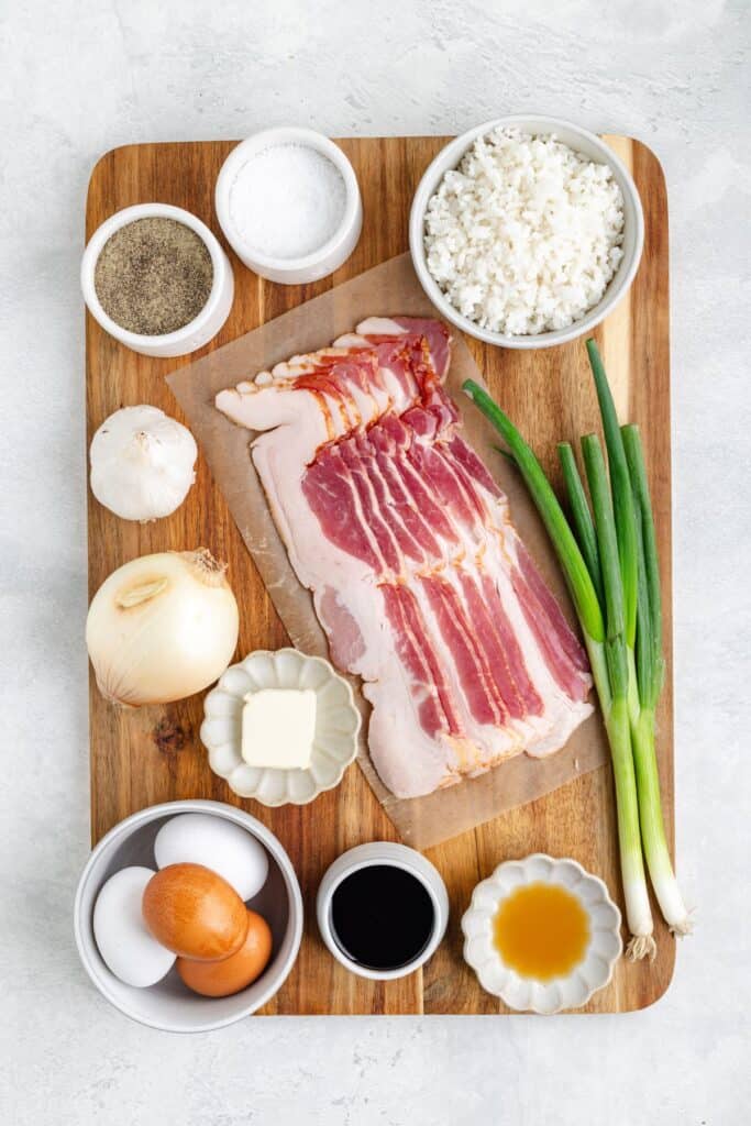 Ingredients on wooden board: sliced bacon, green onions, onion, garlic, eggs, butter, seasoning, rice, soy sauce.