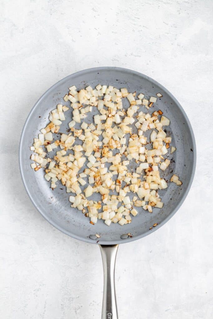 Diced onions sautéing in a frying pan, some pieces golden brown, on a light-colored countertop.