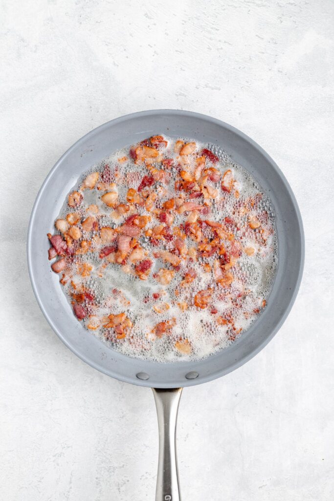 Crisped bacon pieces sizzling in a frying pan, bubbles of rendered fat visible, on light countertop.