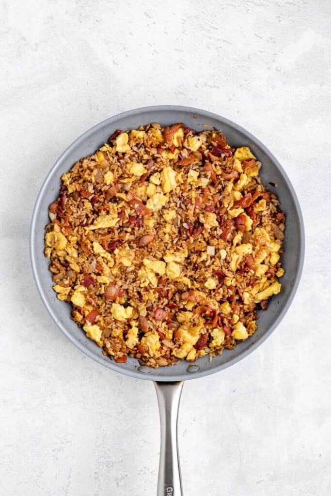 Scrambled eggs, rice, and bacon cooked together in a frying pan on a light gray surface.