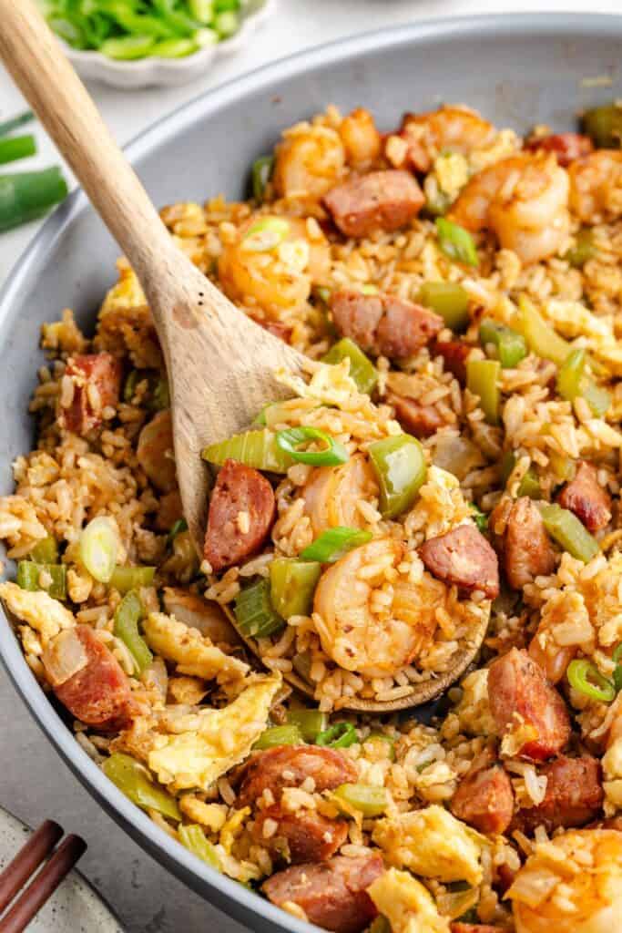 Skillet of cajun shrimp and sausage fried rice with eggs, peppers, and green onions stirred with wooden spoon.