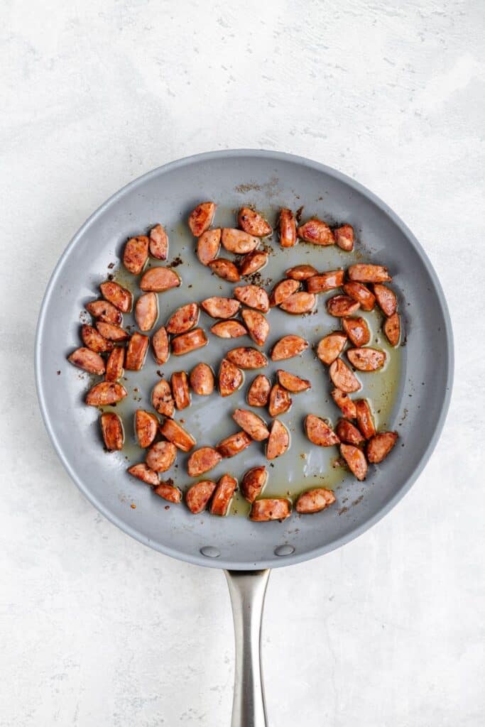 Sliced sausage pieces sautéing in a gray nonstick frying pan with oil.