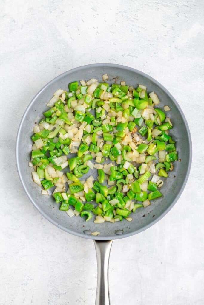 Chopped green bell peppers and onions sizzling in a nonstick frying pan on a light kitchen counter.