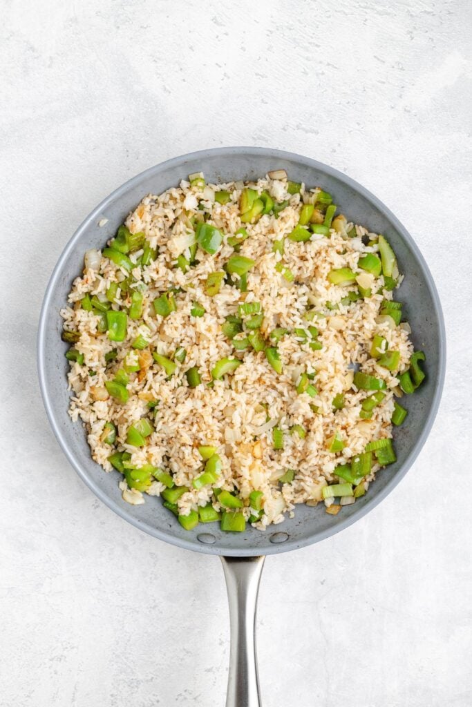 Frying pan with cooked rice, chopped green bell peppers, and onion pieces on a light background.