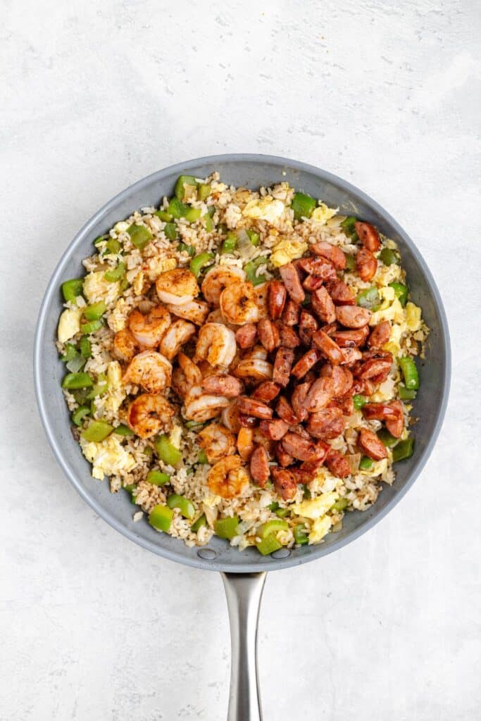 Skillet filled with fried rice, green bell peppers, scrambled eggs, grilled shrimp, and sliced sausage on light background.