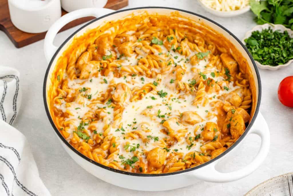 White pot of cheesy enchilada pasta with chicken, tomato sauce, and parsley on a light surface.