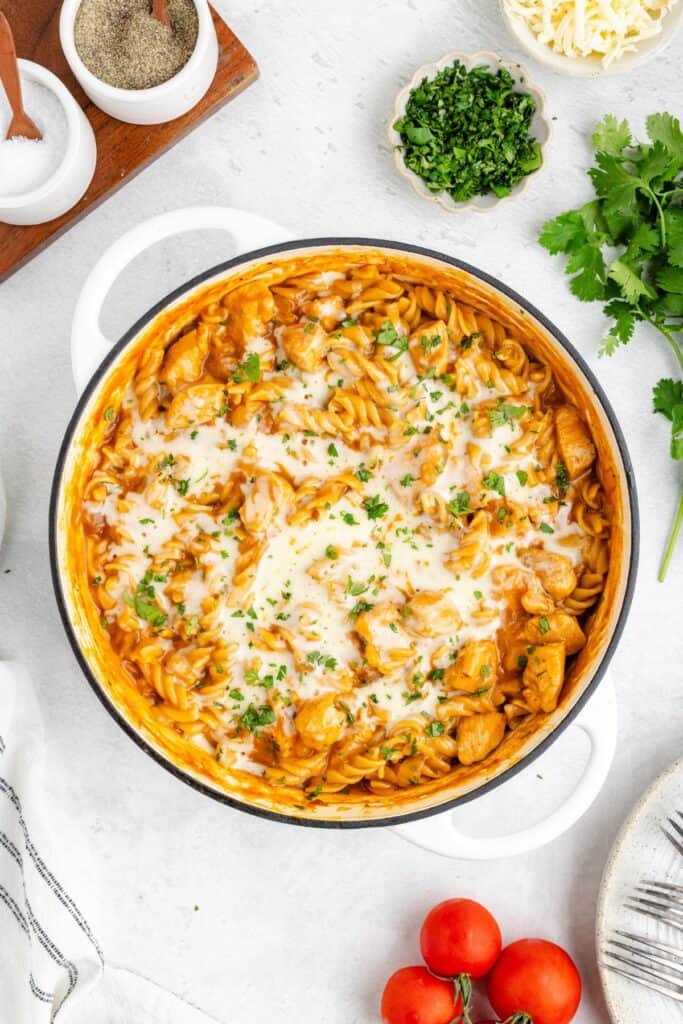 Pot of enchilada chicken pasta with, melted cheese; surrounded by tomatoes, parsley, cheese, salt, pepper.
