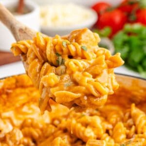 Wooden spoon holds cheesy chicken enchilada pasta with herbs above pot; fresh vegetables blurred in background.