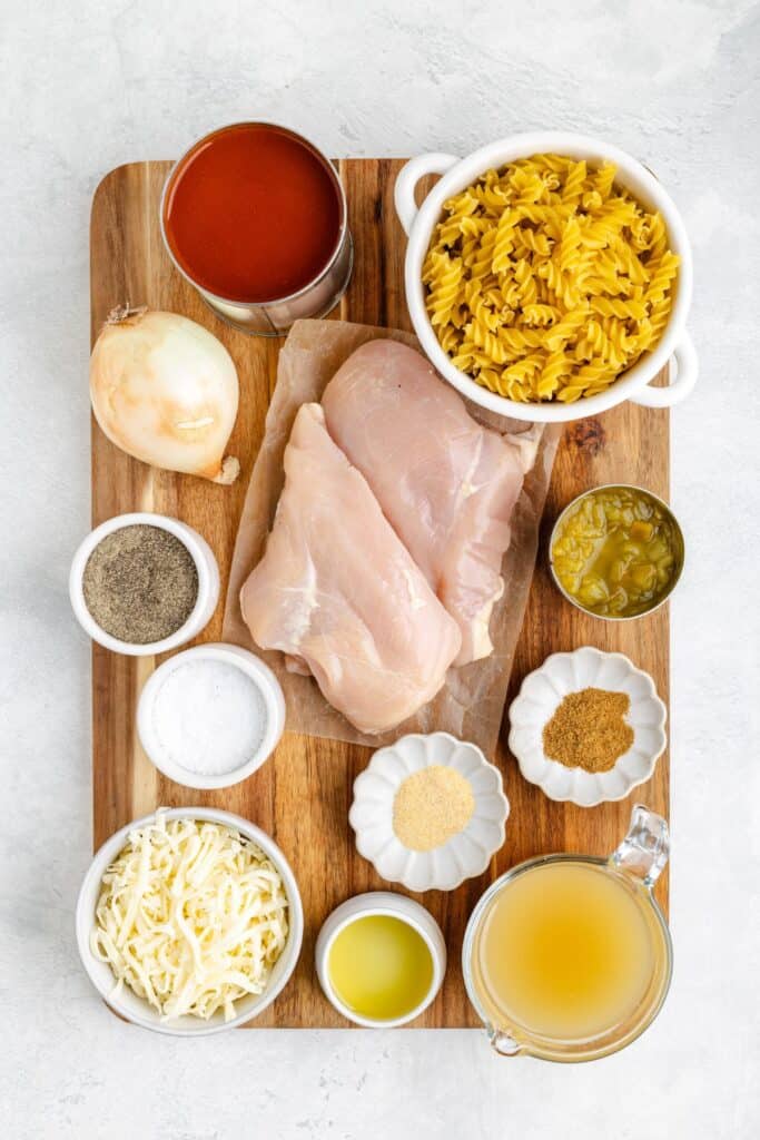 Raw chicken breasts and pasta ingredients arranged on a wooden board, ready for cooking, including spices and vegetables.