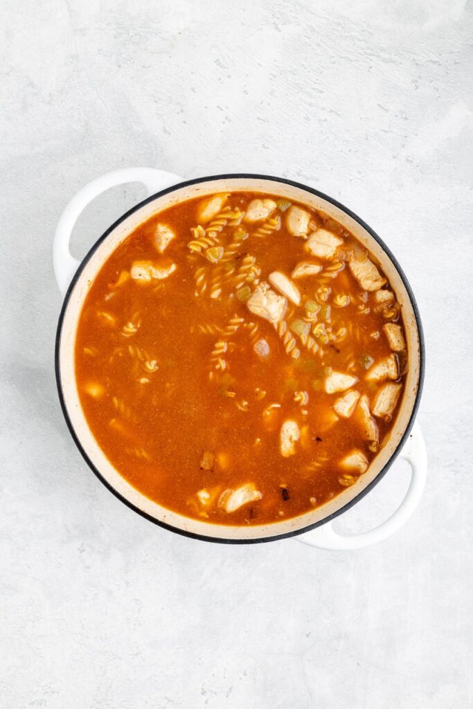 White pot with enchilada sauce, spiral pasta, and chicken chunks sits on a light gray surface.