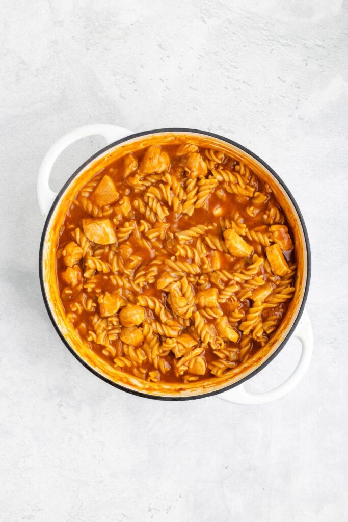 White pot with rotini pasta and meat chunks in red enchilada sauce on a light-colored surface.