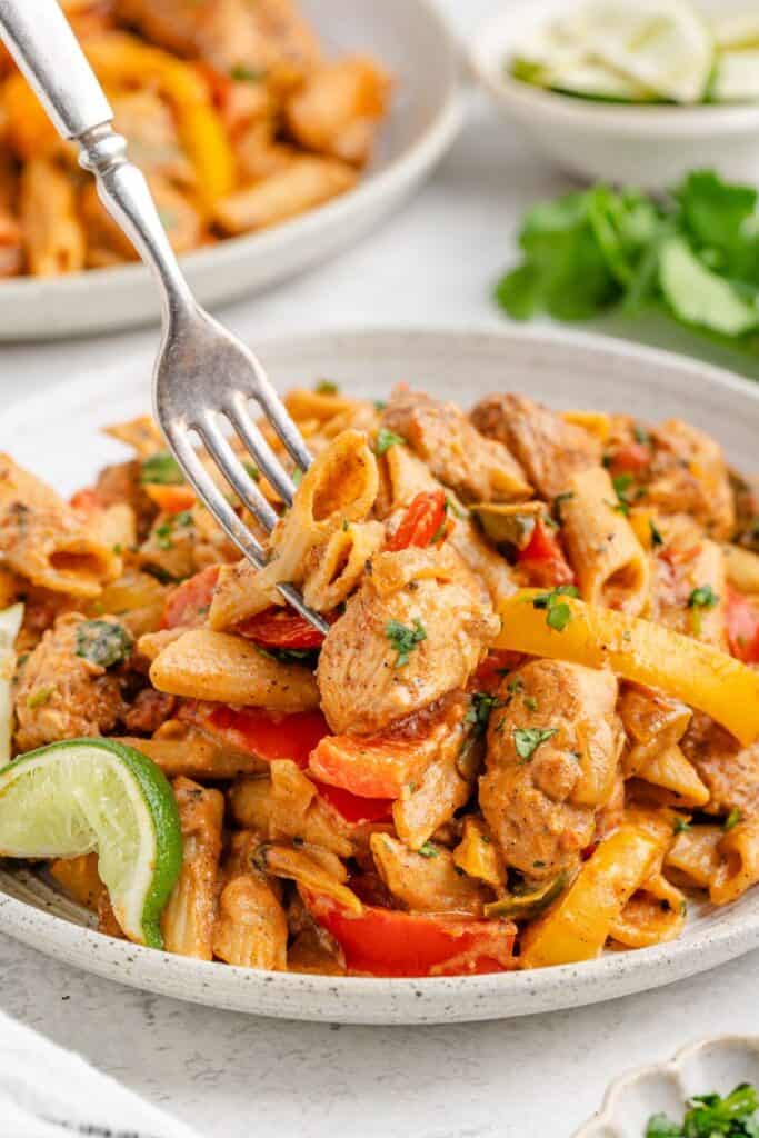 Creamy chicken fajita pasta with bell peppers and herbs, lime wedge garnish; fork lifts a bite from the plate.