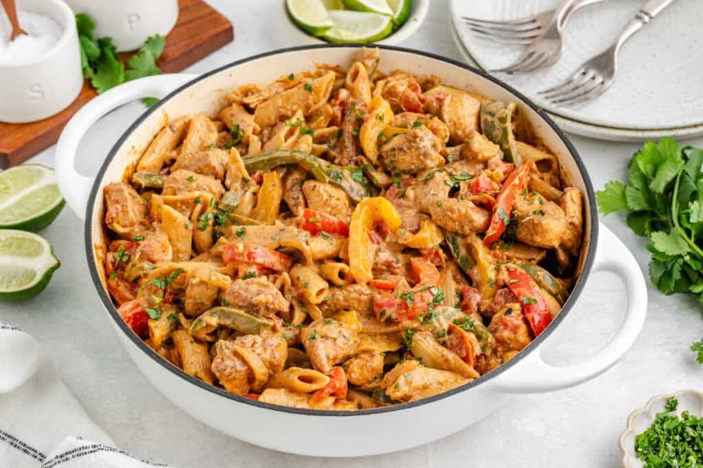 White pot of one pot chicken pasta with bell peppers and herbs, surrounded by lime wedges, plates, and cutlery.