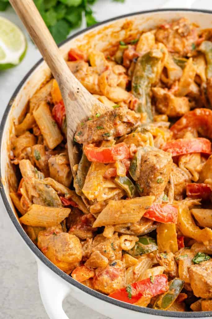 Wooden spoon scooping fajita pasta with peppers and onions from a white pot.