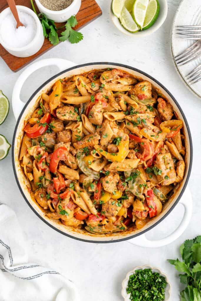 Pot of creamy fajita chicken pasta with bell peppers and herbs, surrounded by plates, lime wedges, salt, and cilantro.