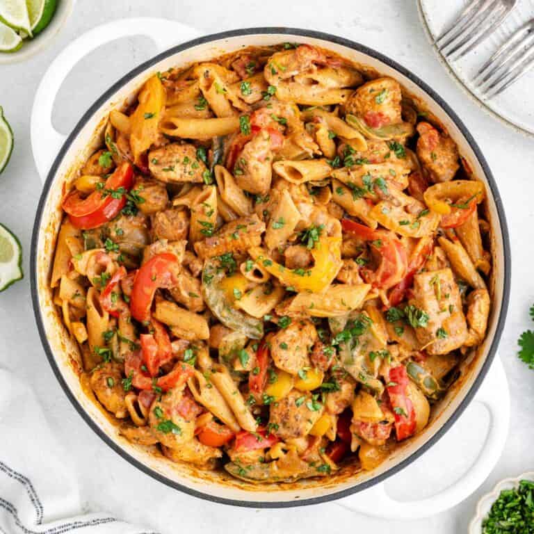 White pot with creamy penne pasta, chicken, bell peppers, and mushrooms; plates and lime wedges nearby.