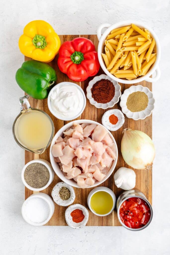 Ingredients on wooden board: chopped chicken, penne, bell peppers, onion, garlic, canned tomatoes, broth, sour cream, spices.