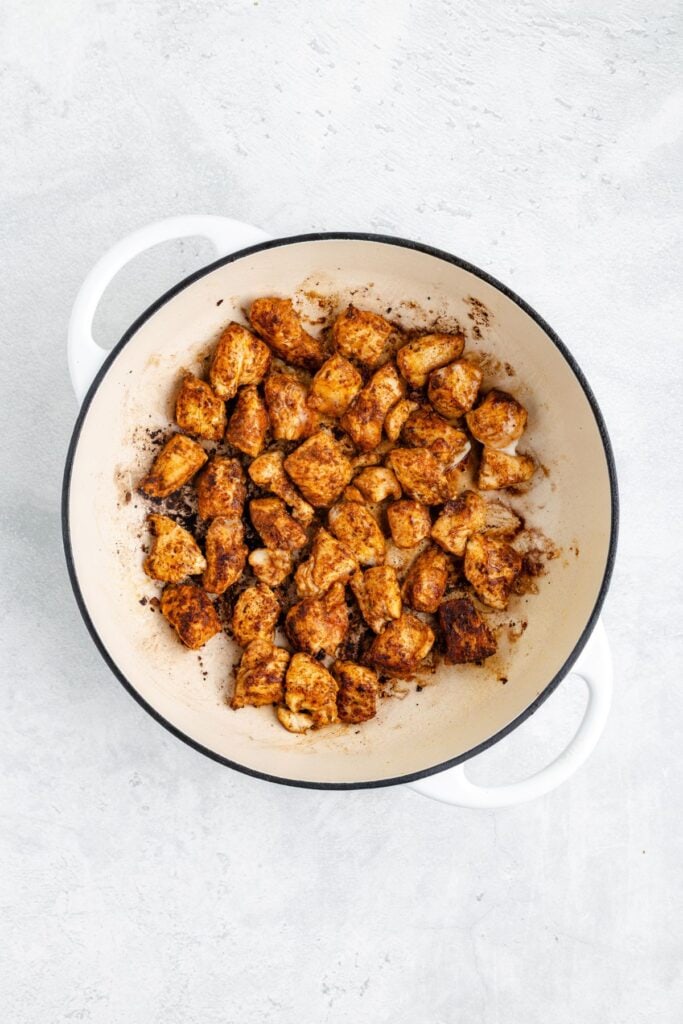 Chunks of browned, seasoned chicken cooking in a white pot with handles on a light textured surface.