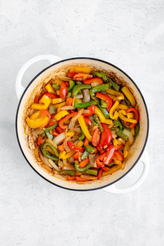 Sliced red, yellow, and green bell peppers with onions sautéing in a white pot on a light surface.