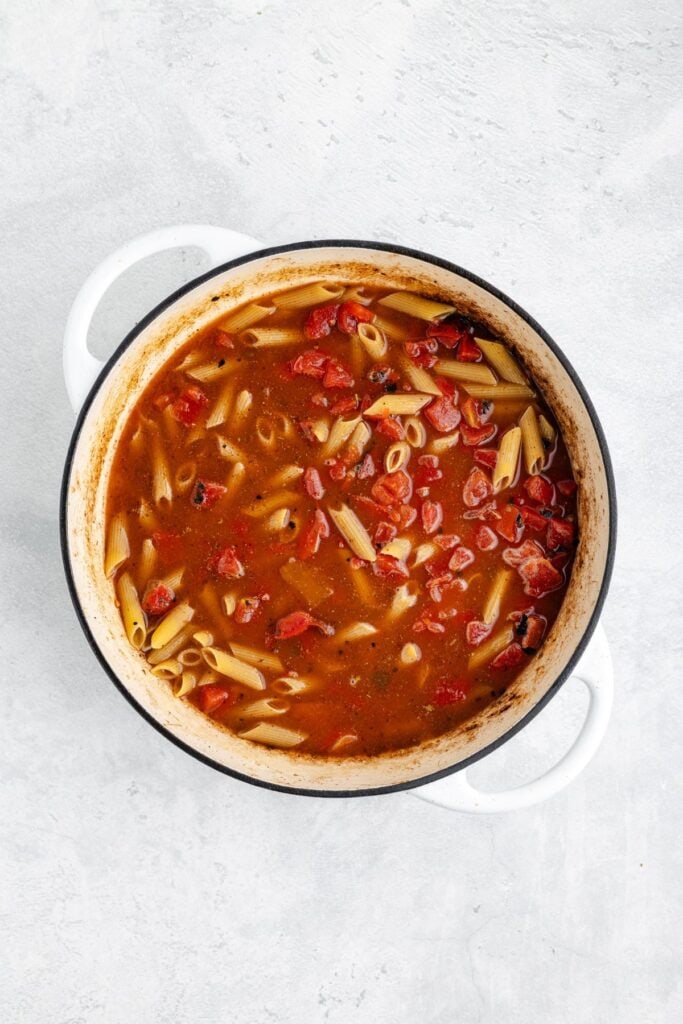 White pot with tomato-based soup, penne pasta, and diced tomatoes, viewed from above on light background.
