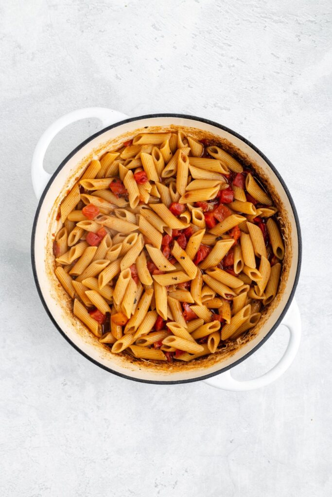 White pot filled with penne pasta and diced tomatoes in sauce on a light-colored surface.
