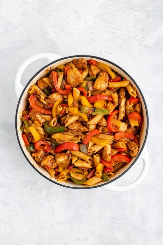 White dish of penne pasta with chicken, red and green bell peppers in sauce on gray background.