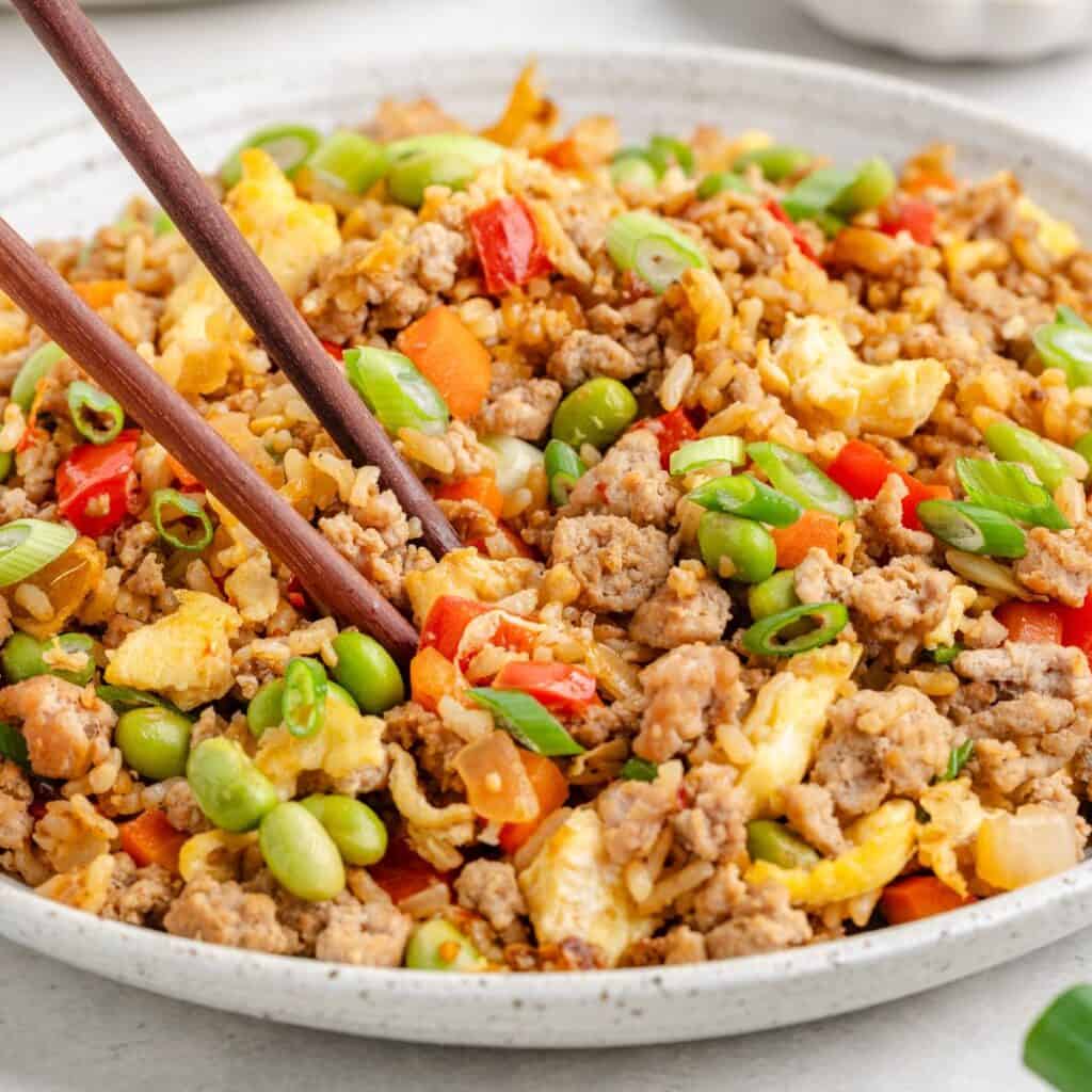 Fried rice with ground meat, eggs, edamame, peppers, green onions; chopsticks lifting a portion.