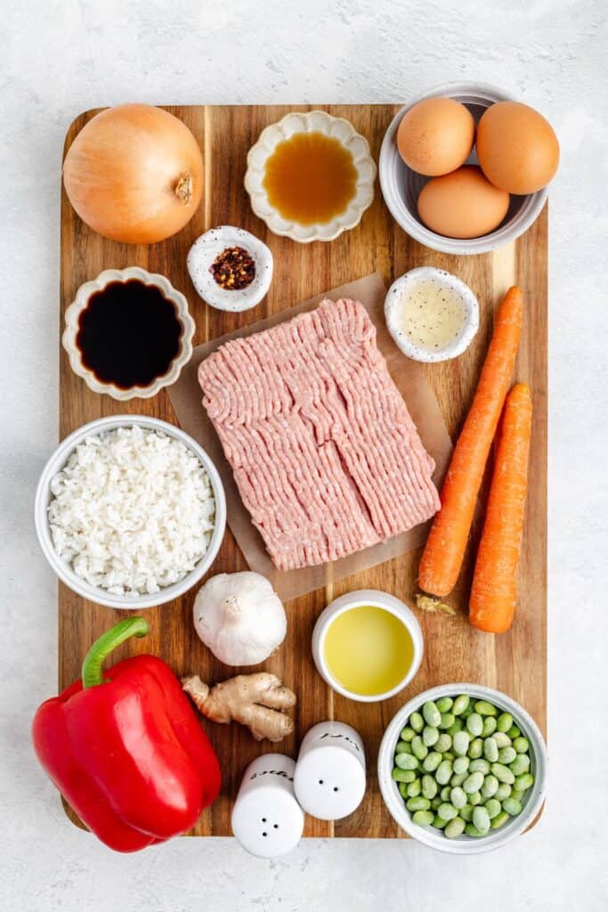 Ingredients for fried rice, including ground turkey, eggs, vegetables, cooked rice, seasonings, and sauces on a wooden board.