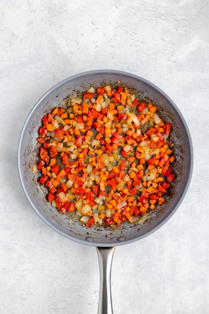 Sautéed diced red bell peppers, onions, and carrots in a metal frying pan on a light surface.